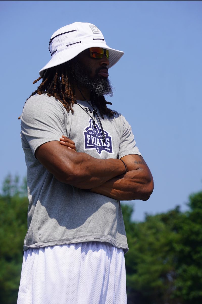 Coach Dante Coles standing on the field with his arms crossed, wearing a bucket hat